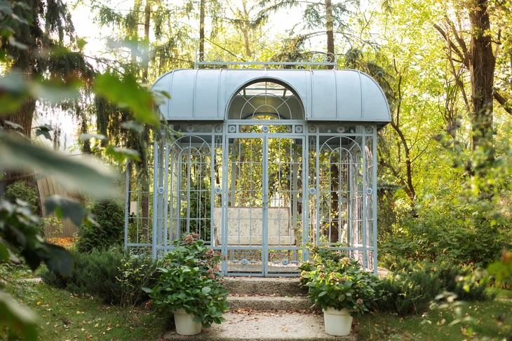 Scène extérieure dans jardin avec serre victorienne ornementale en métal peint bleu-gris, structure arquée avec verrières et portes vitrées ouvertes