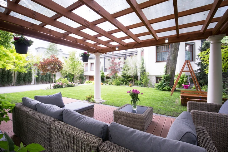 Terrasse couverte avec pergola en bois et colonnes blanches, mobilier de jardin en résine tressée avec coussins gris, donnant sur pelouse et aire de jeux