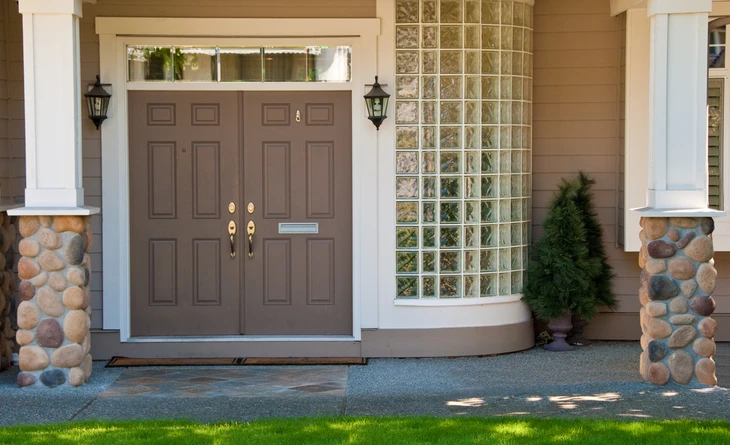 Entrée principale d'une maison résidentielle avec double porte en bois brun à panneaux surélevés, encadrée par des colonnes blanches cannelées sur bases en pierre naturelle multicolore