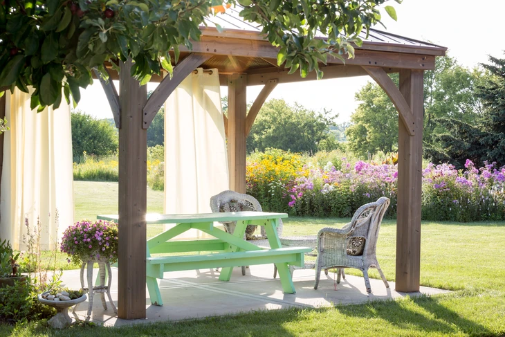 Scène extérieure de jardin avec pergola en bois abritant une table de pique-nique verte et chaises en rotin tressé