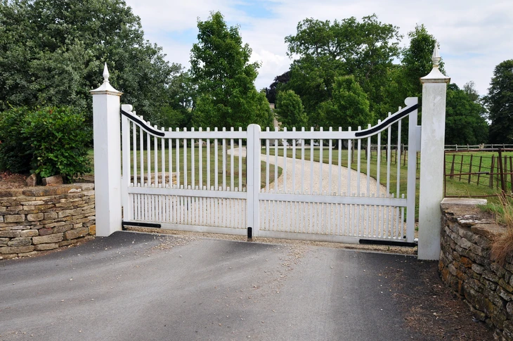 Entrée de propriété avec portail automatique blanc à double battant en PVC ou aluminium laqué, piliers blancs à chapiteaux pyramidaux