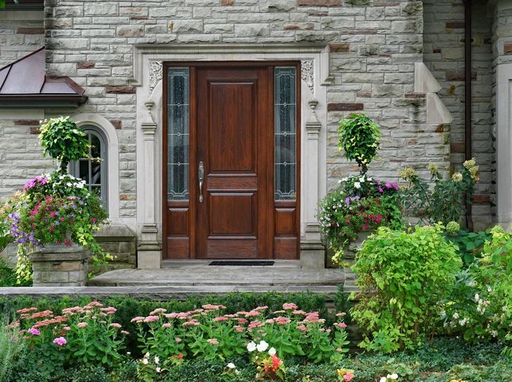 Entrée principale d'une maison en pierre naturelle avec porte en bois massif à panneaux sculptés, encadrée de colonnes décoratives et vitraux latéraux
