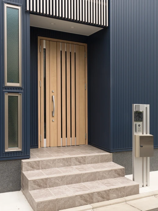Entrée moderne d'une maison contemporaine avec porte d'entrée en bois clair à lames verticales, escalier en pierre naturelle beige, façade en bardage métallique bleu foncé nervuré