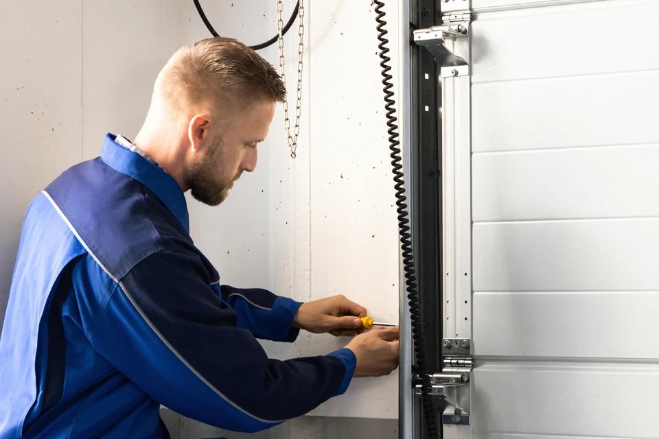Technicien en vêtements professionnels mesurant ou inspectant l'encadrement d'une porte de garage lors d'une visite de chantier chez un client. La porte de garage doit être clairement visible en arrière-plan, l'artisan doit être en train de prendre des mesures ou d'évaluer le projet, illustrant ainsi l'accompagnement personnalisé et le suivi du chantier qui caractérisent le service de DNS Fenêtres.